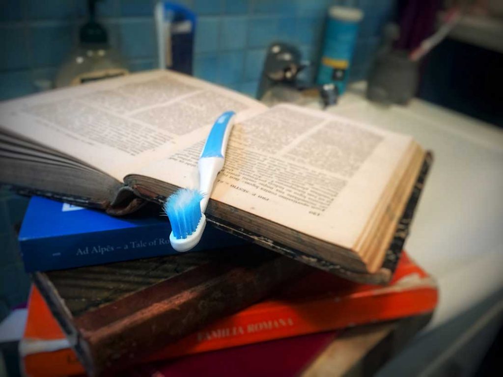 Latin daily habit illustrated by a toothbrush laying on top of a pile of Latin books, the top one open.